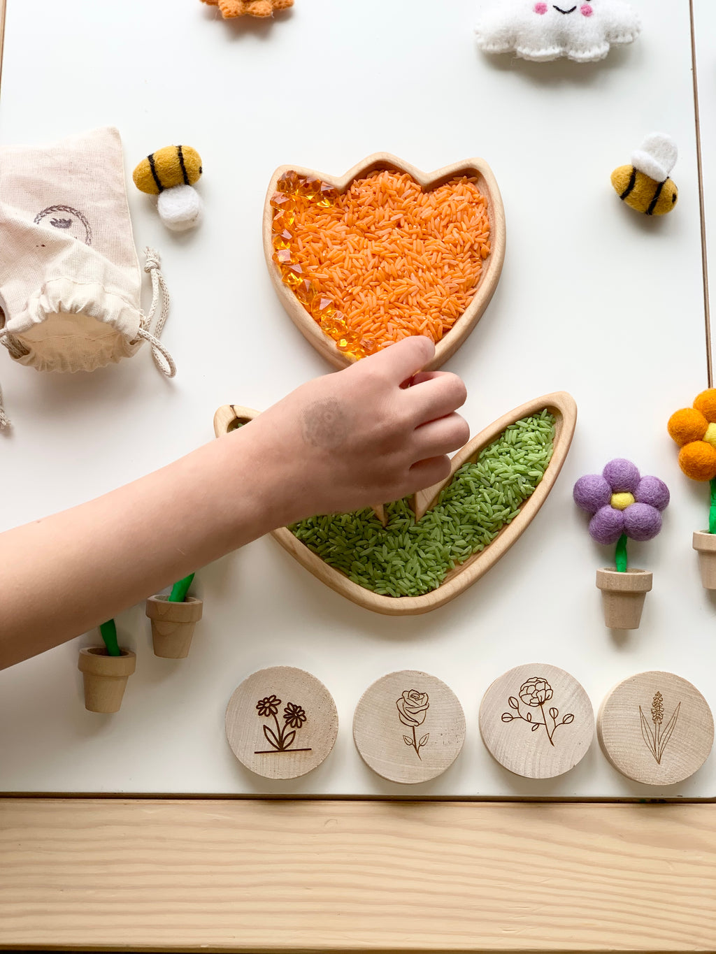 Child's hand touching two wooden tulip flower plates filled with colored rice on white surface with wooden flower tokens and felt bee toys