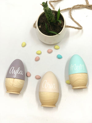 Three personalized hollow wooden Easter eggs in pastel purple, cream, and blue with white script names on white background with scattered candies and small plant
