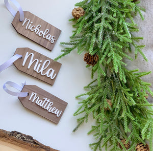 Custom 2x4 inch wooden Christmas stocking tags with white cursive names and white ribbon near green pine branches with pinecones