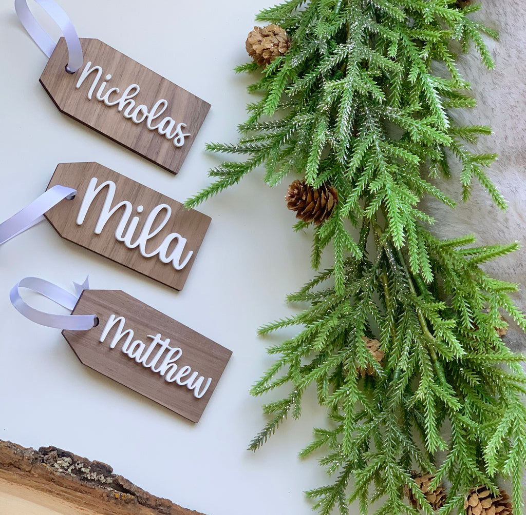 Custom 2x4 inch wooden Christmas stocking tags with white cursive names and white ribbon near green pine branches with pinecones