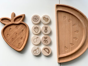 Natural wood 2-inch discs with engraved fruit illustrations arranged between strawberry and watermelon shaped wooden trays on white background