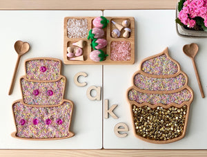 Wooden sensory trays shaped like a layered cake and cupcake filled with colorful sensory materials on white surface