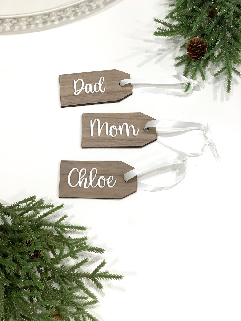 Set of three custom 2x4 inch wooden gift tags with white cursive names and white ribbon on white background with pine decor