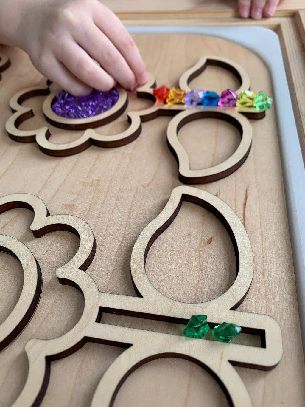 Child's hand placing colorful acrylic treasure gems into flower-shaped wooden puzzle on natural wood background