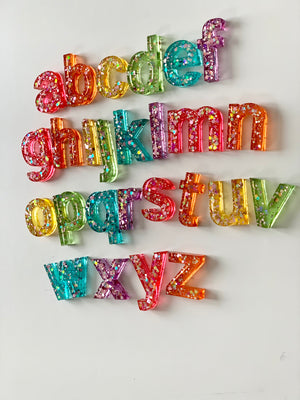 Rainbow glitter resin lowercase alphabet letters arranged in rows on white surface