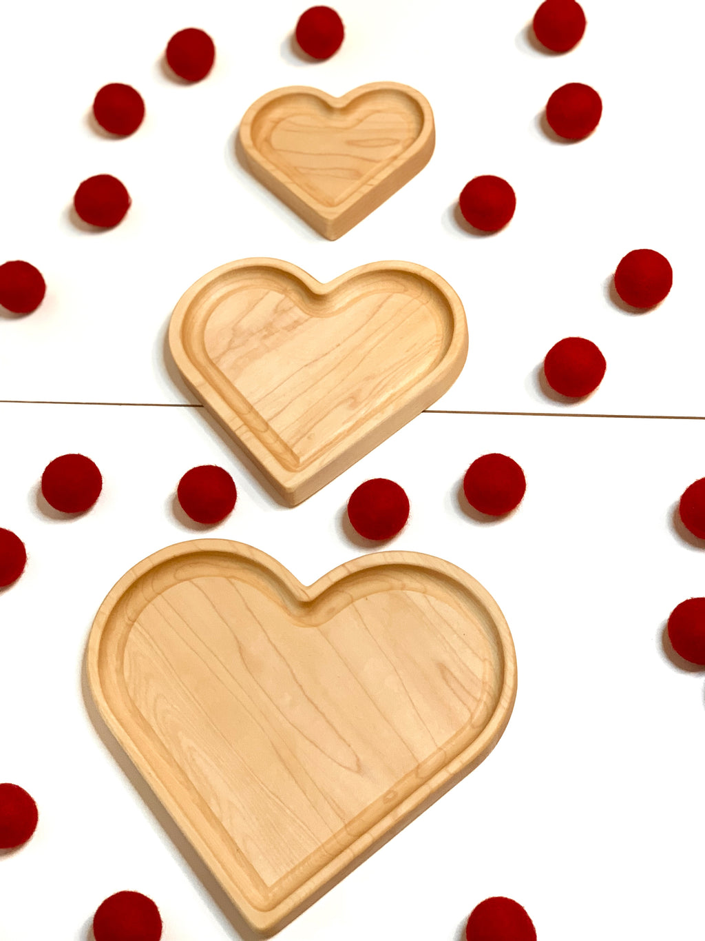 Three natural wooden nesting heart-shaped plates arranged vertically with red pom-poms on white background