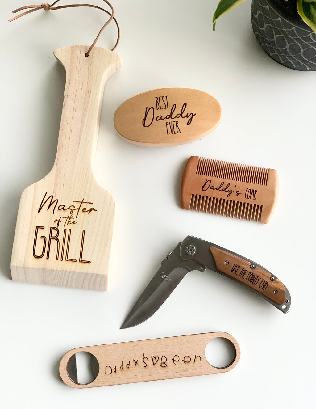 Set of personalized wooden and metal gifts including a paddle, comb, bottle opener, and pocket knife with engraved text for dads