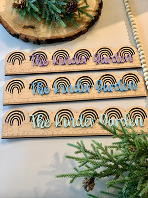 Three personalized wooden rainbow rulers with "The Kinder Garden" in purple, blue, and light blue on natural wood background