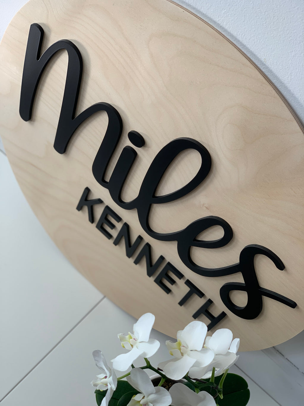 Oval natural wood 3D round name sign with black cursive and block letters displayed near white orchids