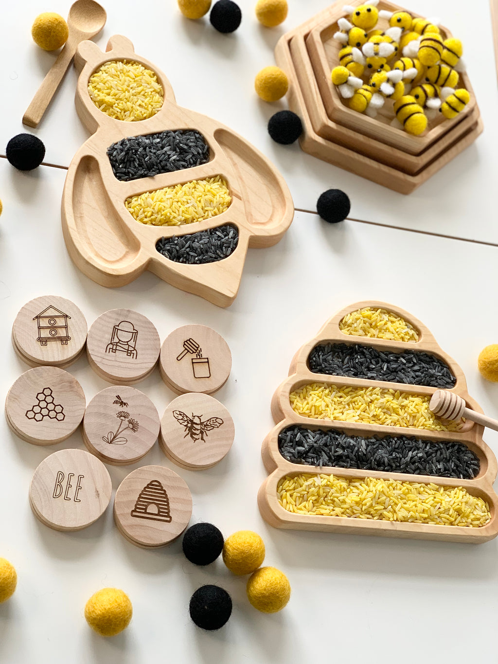 Wooden bee-shaped sensory trays filled with yellow and black rice, wooden stamps with bee designs, and felt balls scattered around