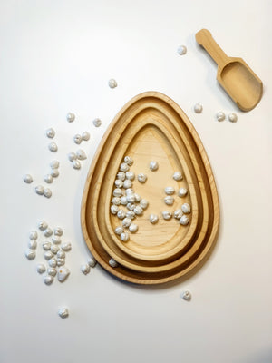 Set of three natural wooden nesting egg plates with white beads and a small wooden scoop on white background