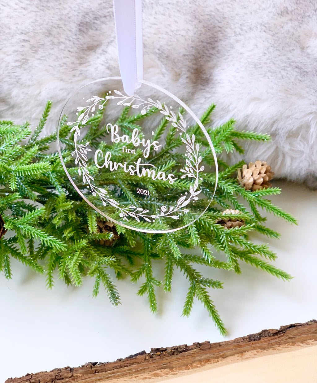Round clear acrylic baby’s first Christmas ornament engraved with white floral wreath and white ribbon on greenery background