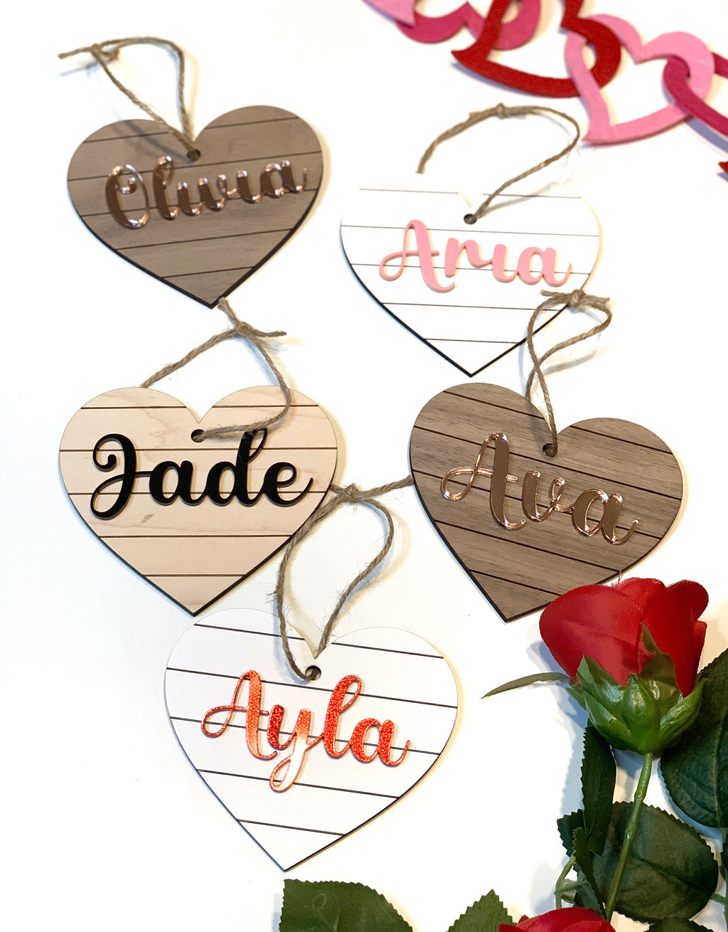 Heart shaped wooden name tags with striped backgrounds and personalized script names hanging on twine with a red rose nearby