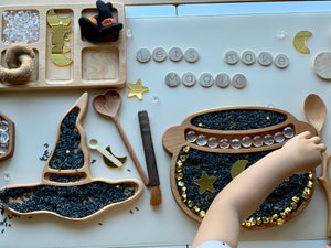 Wooden witch hat and cauldron sensory trays filled with black rice and gold star decorations on white surface with child’s hand
