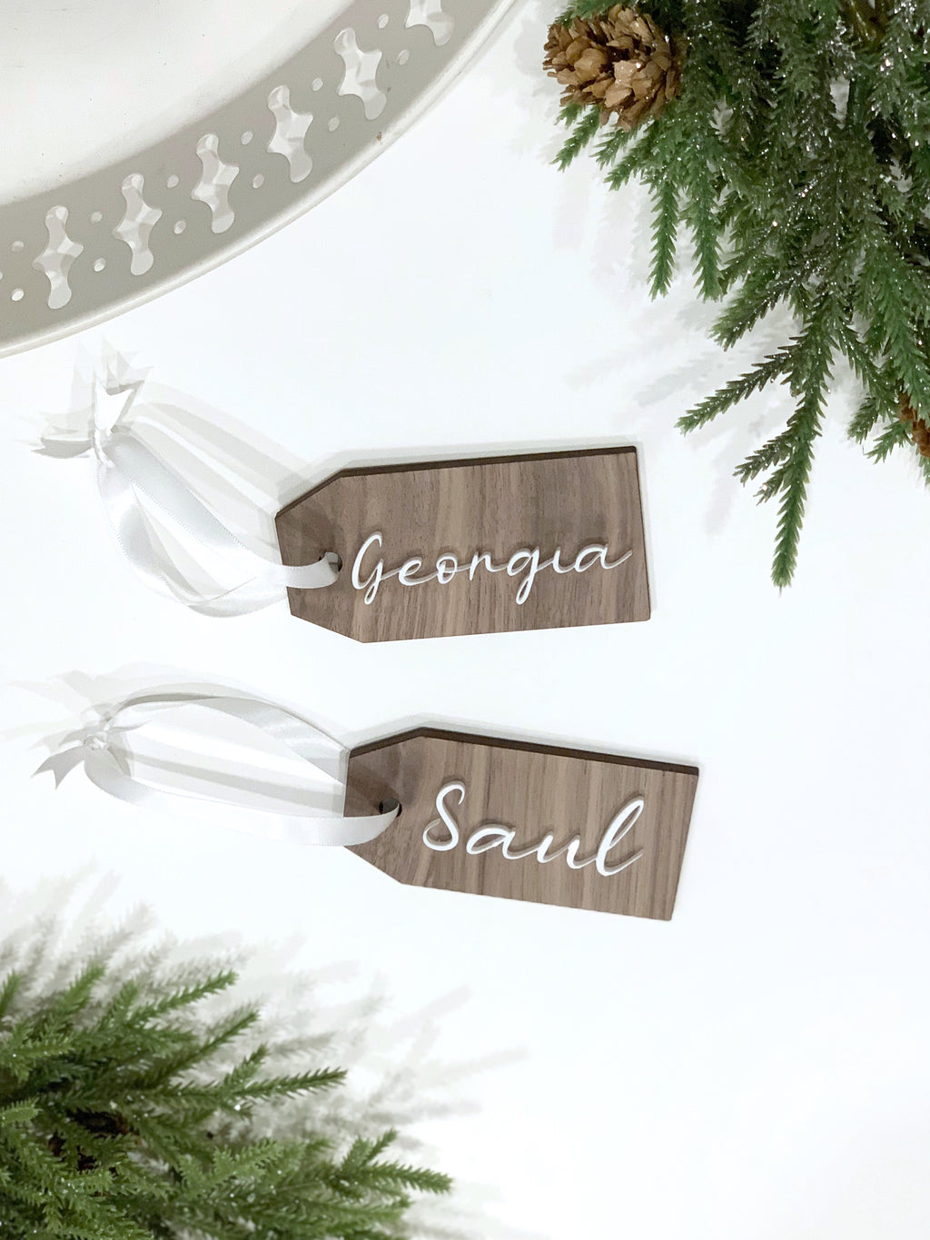 Two custom wooden gift tags with white cursive names on natural wood and sheer white ribbon on white background