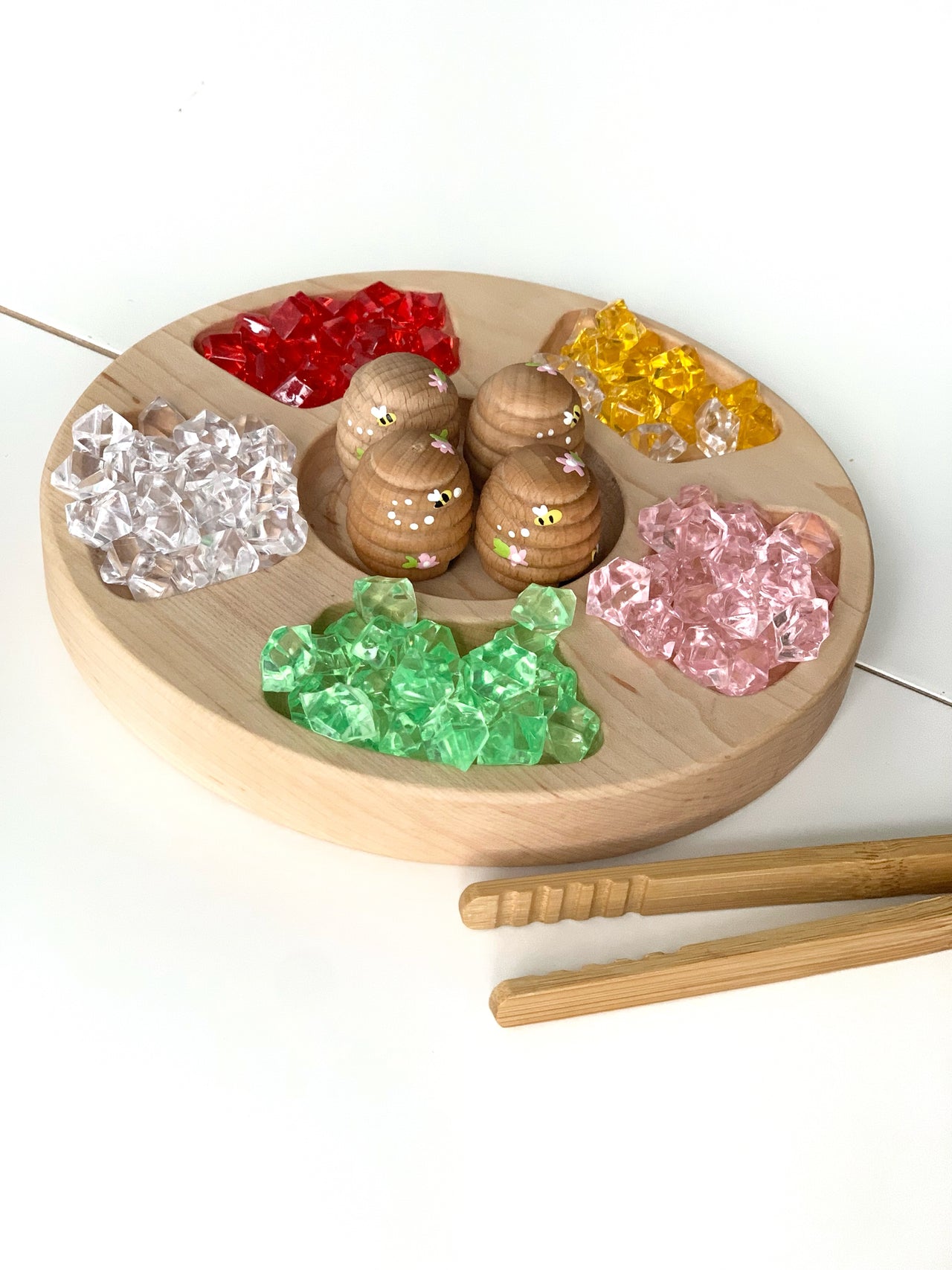 Round wooden tray with colorful acrylic treasure gems in compartments and wooden tongs on white surface
