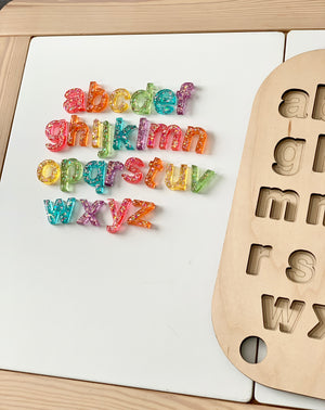 Rainbow glitter resin lowercase alphabet letters arranged next to wooden letter tray on white surface