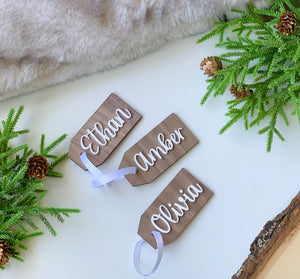 Custom wooden 2x4 inch Christmas stocking gift tags with white cursive names and white ribbon on white background with greenery and pinecones
