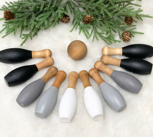 Monochrome wooden 10-piece bowling set with black, gray, and white pins arranged around a wooden ball on white surface with pine branches