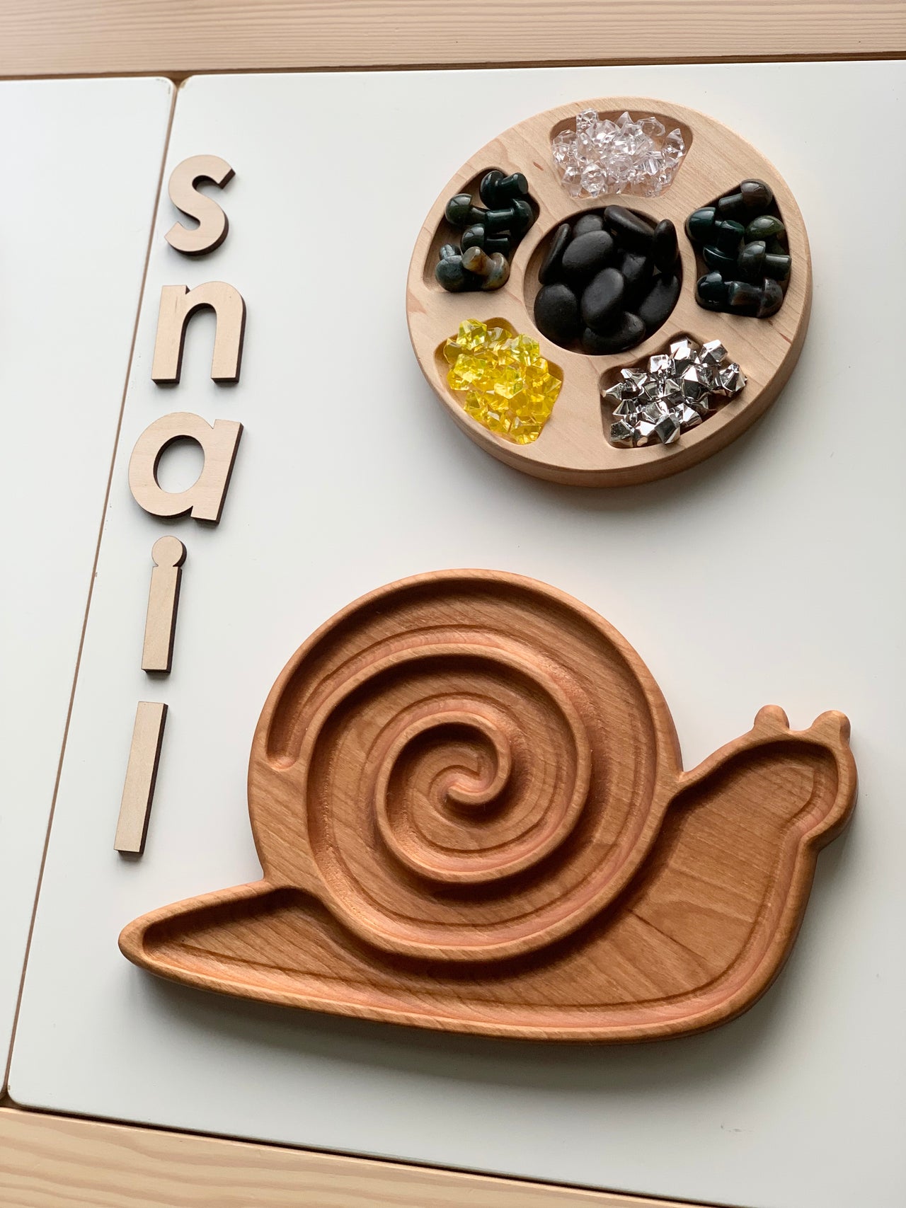 Cherry wood snail-shaped sensory tray with spiral groove beside natural wooden letters spelling snail on white surface