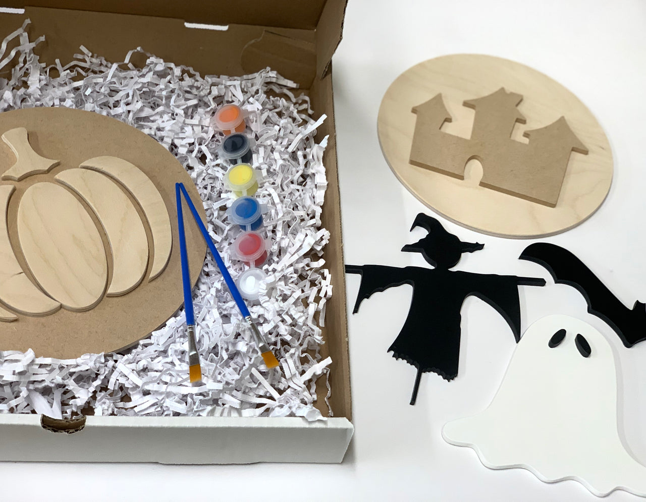 DIY Halloween sign kit with unfinished wooden shapes, paint pots, and brushes in shredded packing material inside a box