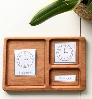 Hardwood three-part card tray with Montessori clock cards showing 3 o’clock on a white surface with plant leaves