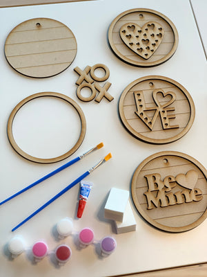 Unpainted wooden circular signs with love-themed designs, paintbrushes, paint pots, and sponges on white surface
