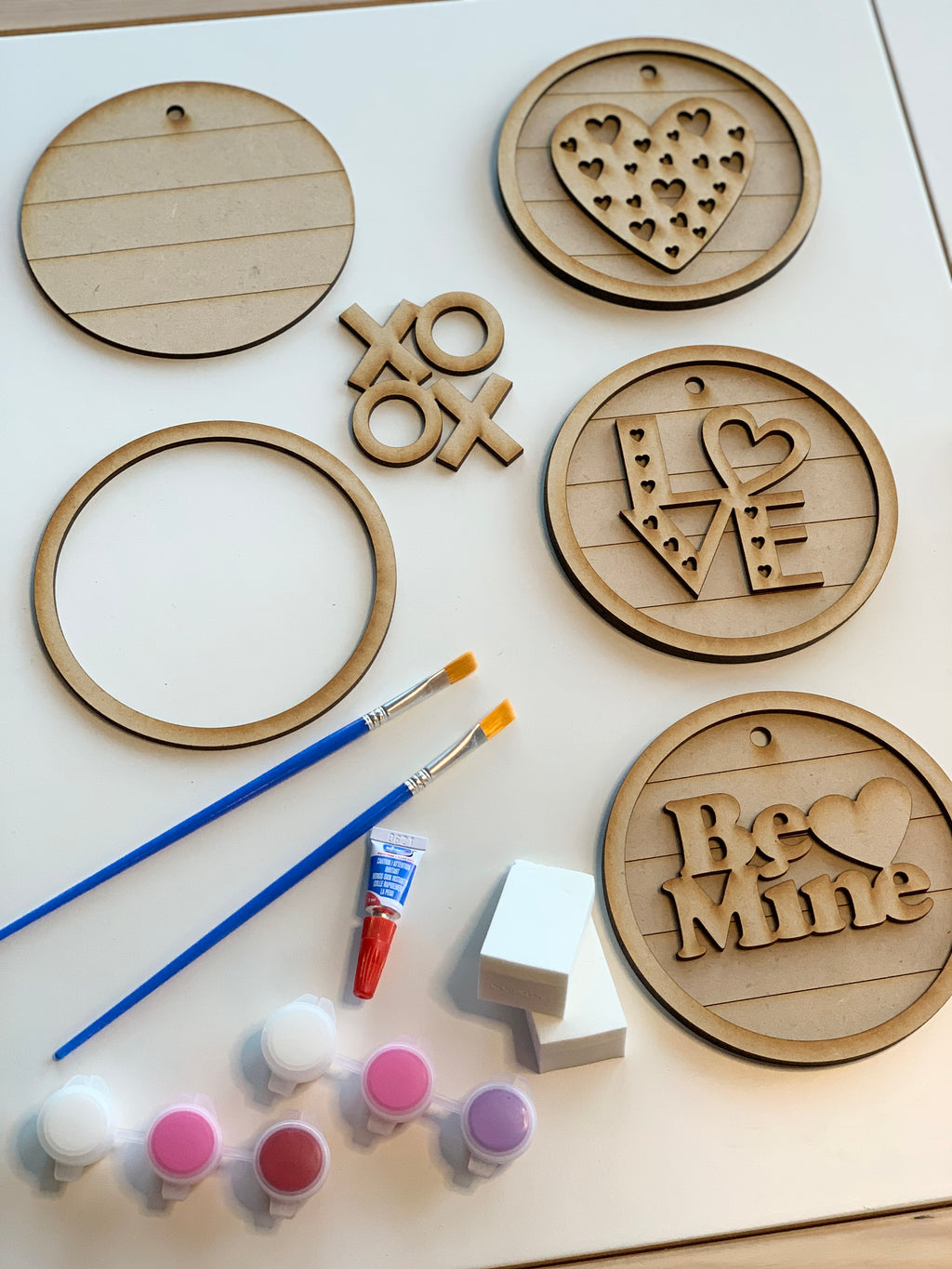 Unpainted wooden circular signs with love-themed designs, paintbrushes, paint pots, and sponges on white surface