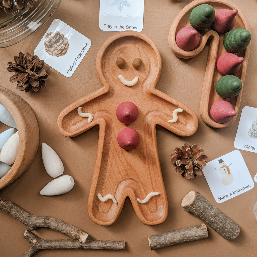 Natural wooden person-shaped plate with pink and white clay decorations surrounded by pine cones, cards, wooden shapes, and sticks