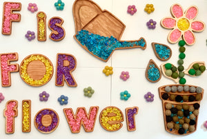 Wooden flower pot sensory tray with felt flowers, green felt balls, watering can tray filled with blue beads, and colorful letter tiles spelling F is for flower