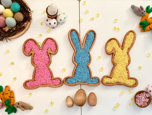 Three wooden bunny-shaped sensory trays filled with pink, blue, and yellow rice surrounded by Easter-themed decorations