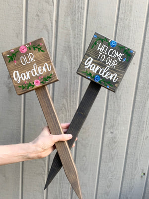 Two small square wooden garden signs with white lettering and floral accents on wood stakes held by a hand