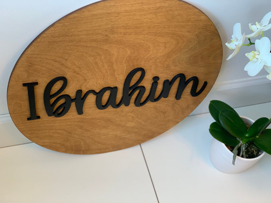 Oval natural wooden 3D round name sign with black cursive letters spelling Ibrahim on white surface near potted plants