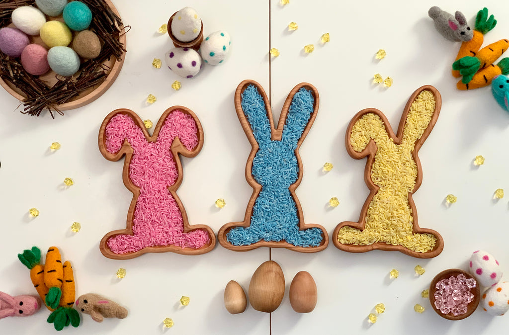 Three wooden bunny-shaped sensory trays filled with pink, blue, and yellow sensory material surrounded by Easter decorations