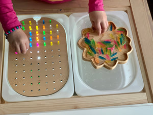 Child playing with peg board insert featuring colorful light pegs and small wooden tray with translucent sticks on wood table