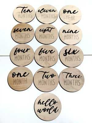 Natural wooden 3D monthly milestone and hello world discs with black cursive and block lettering on light wood background