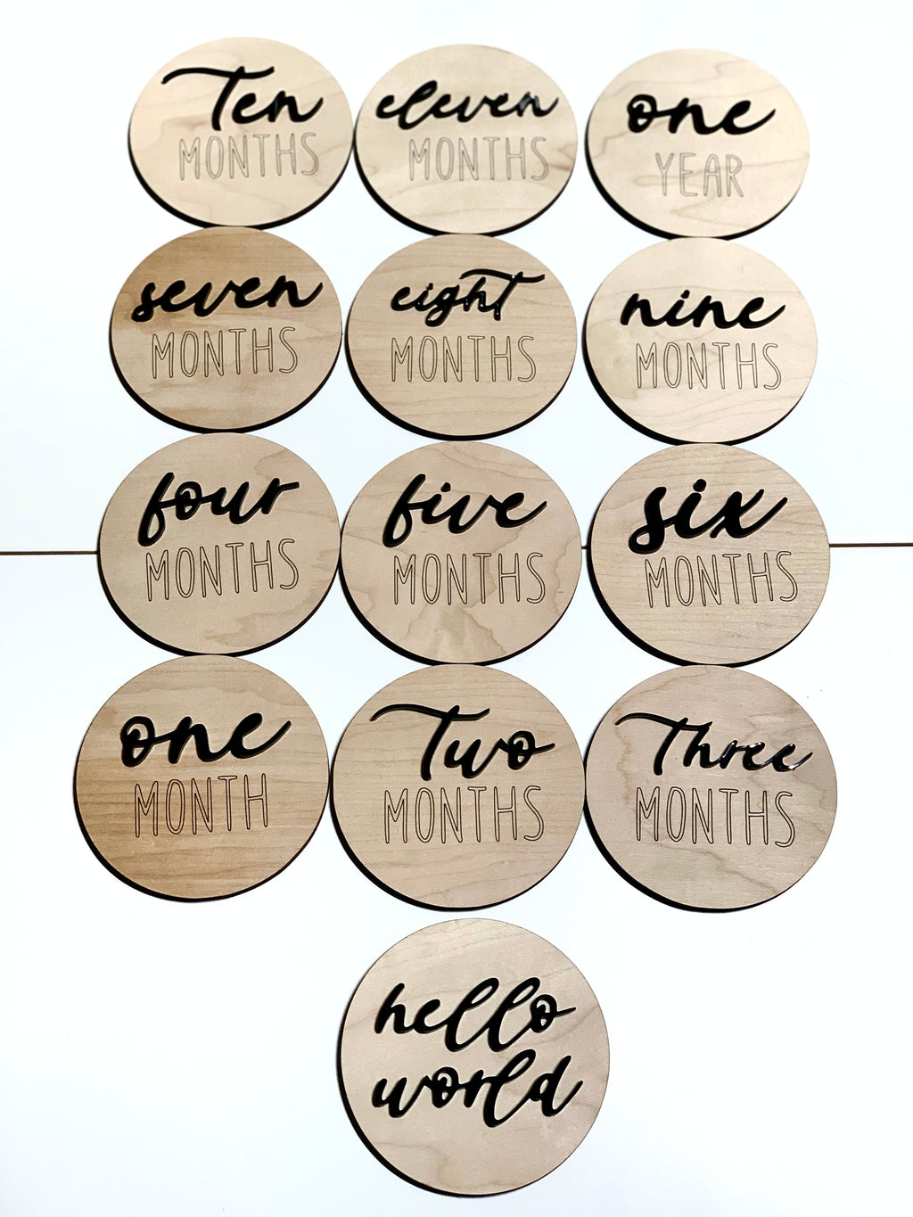 Natural wooden 3D monthly milestone and hello world discs with black cursive and block lettering on light wood background
