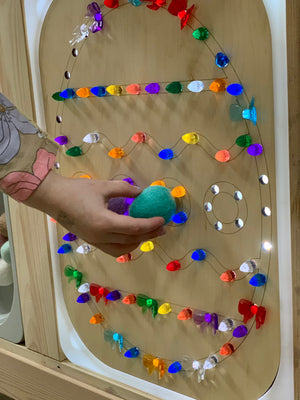 Wooden Easter egg-shaped Flisat insert with colorful light pegs and a child’s hand holding a green felt ball against light wood background