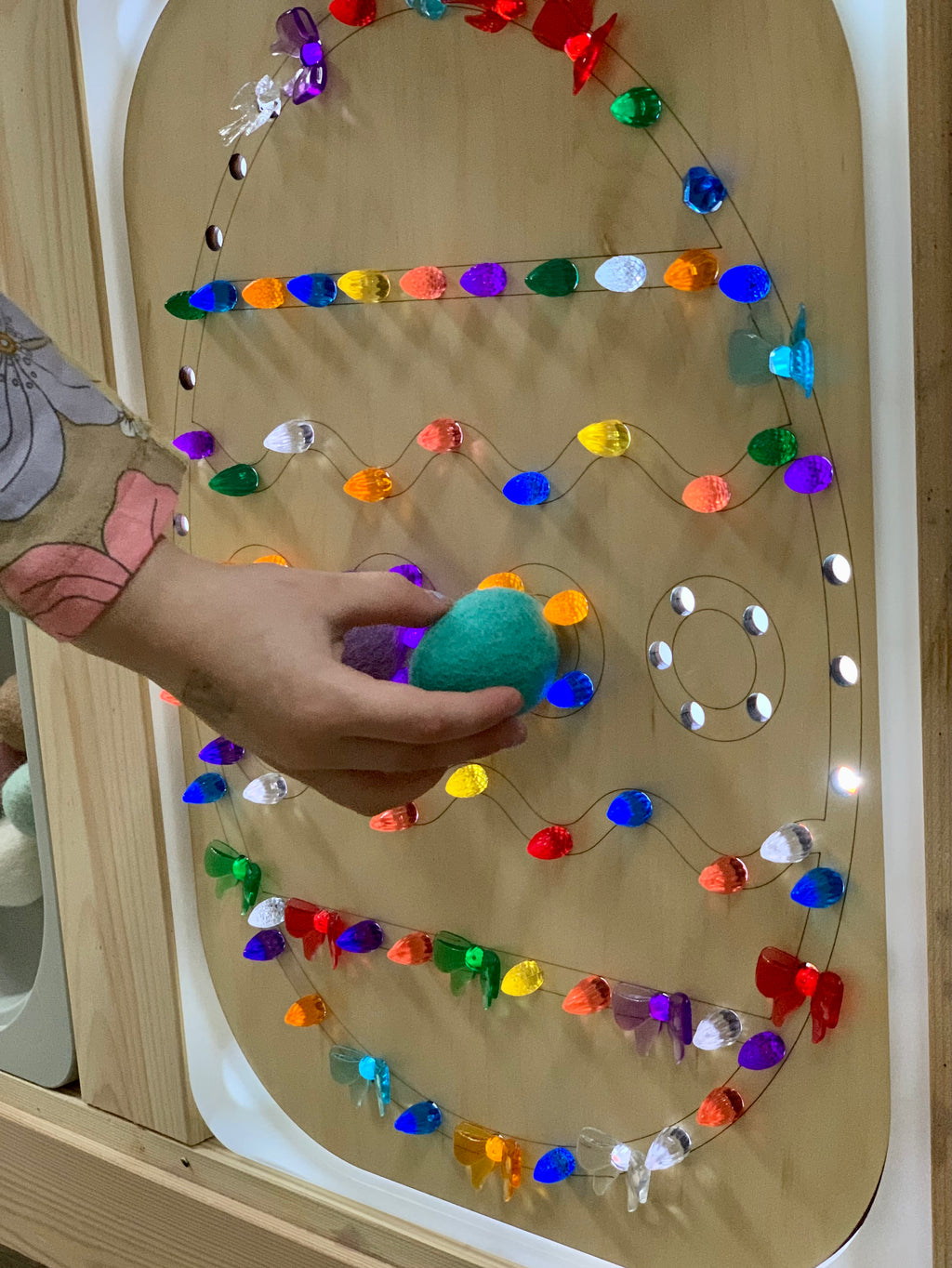 Wooden Easter egg-shaped Flisat insert with colorful light pegs and a child’s hand holding a green felt ball against light wood background