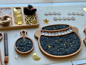 Wooden sensory play trays filled with black dyed rice and gold star confetti with wooden magic-themed accessories