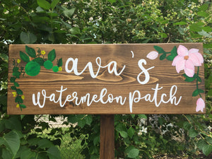 Custom wooden garden sign painted brown with white cursive letters and pink floral and green leaf illustrations on wood grain background