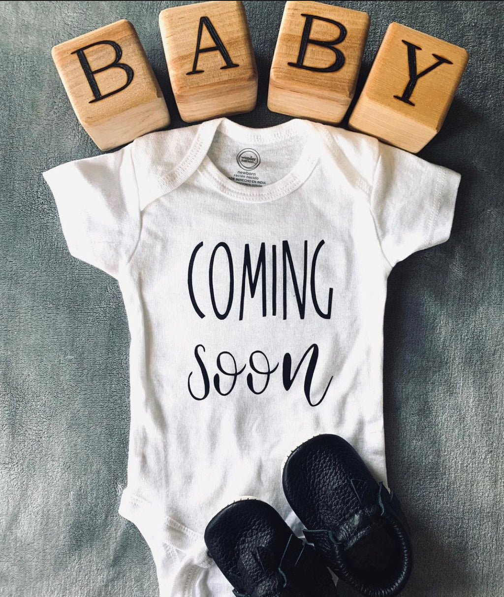 Custom wood blocks spelling BABY above a white onesie with Coming Soon text and black baby shoes on gray background