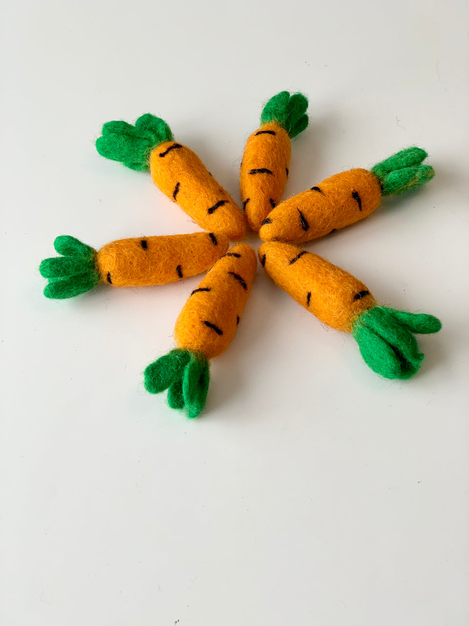 Six small handmade felt carrots with green tops arranged in a circle on white background