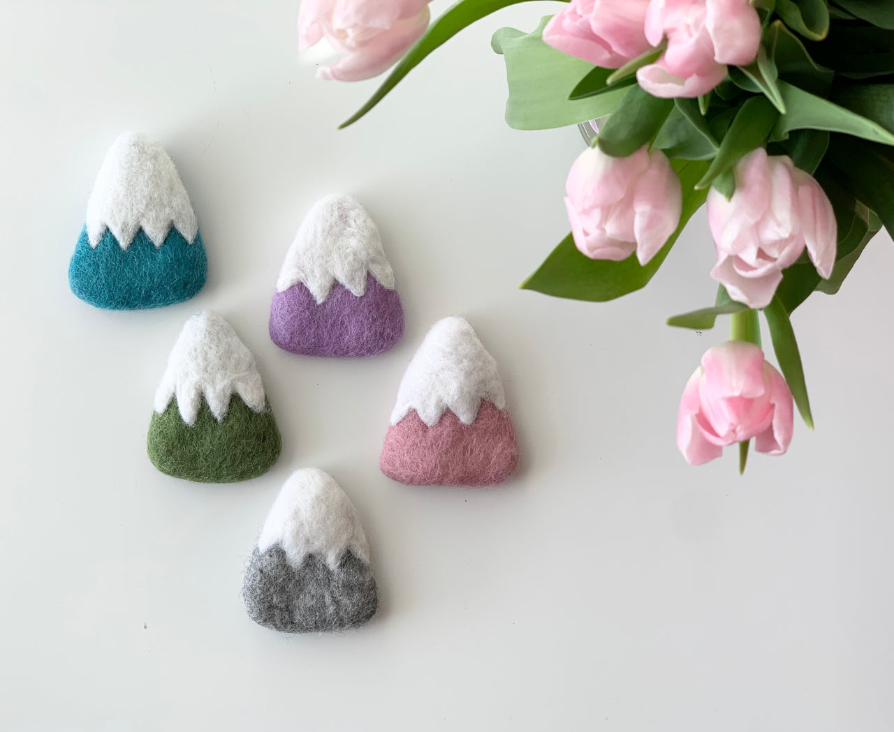 Five handmade felt mountains in blue, purple, green, pink, and gray with white snowy tops on white background near pink tulips