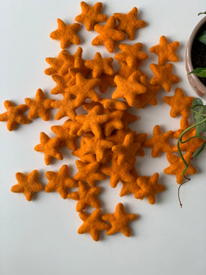 Multiple small bright orange handmade felt stars scattered on white surface near potted green plant