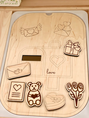 Wooden love post mailbox tabletop insert with engraved outlines and six small wooden Valentine-themed shapes on light wood surface