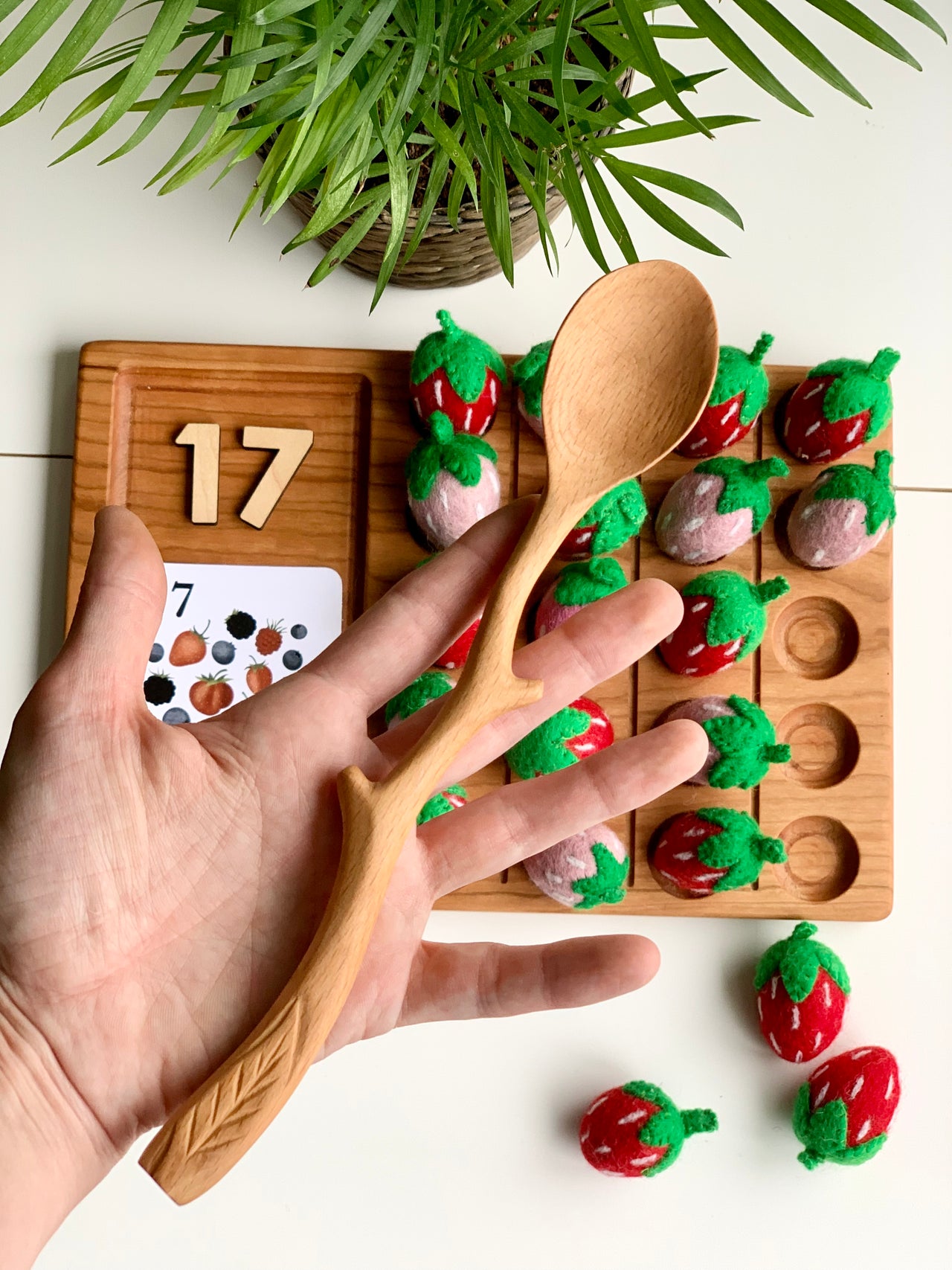 Hand holding a natural wood spoon shaped like a twig with carved leaf details over a wooden toy and green plant background