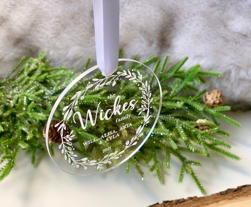 Round clear acrylic Christmas ornament engraved with family names and decorative leafy wreath hanging on white ribbon
