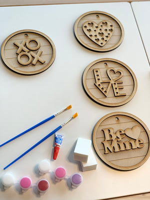 Four unfinished wooden love-themed DIY hanging signs with paints, brushes, and sponges on white surface