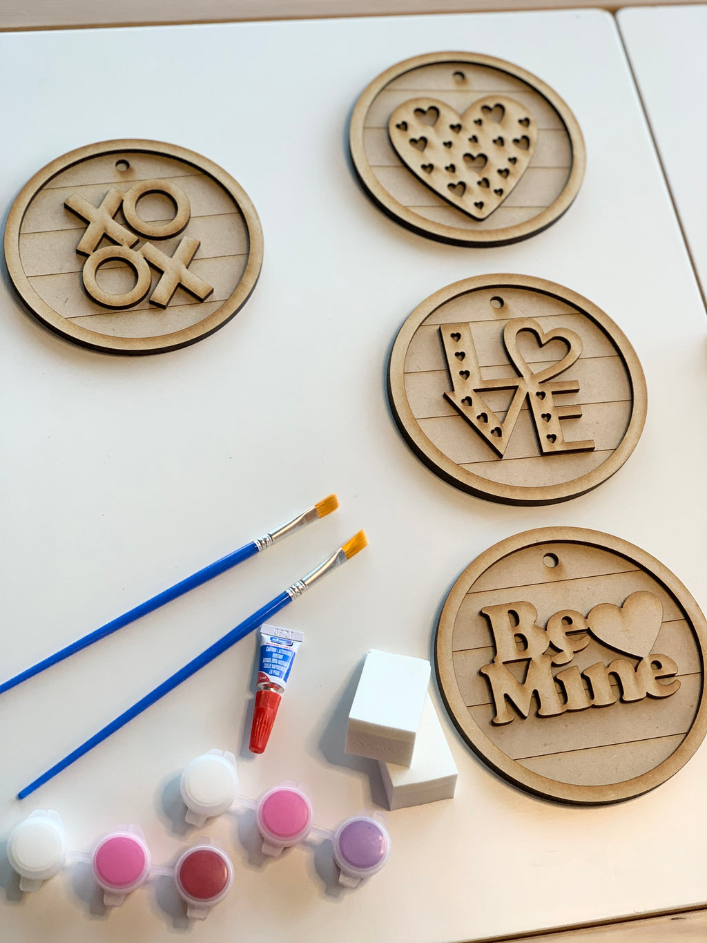 Four unfinished wooden love-themed DIY hanging signs with paints, brushes, and sponges on white surface